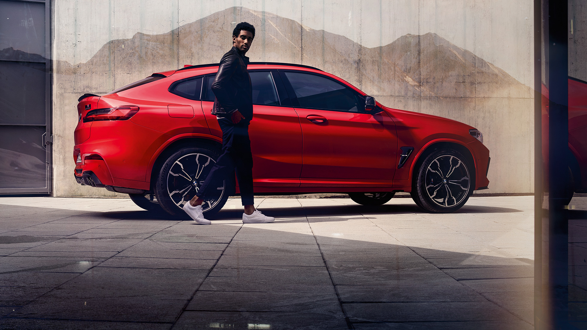 BMW X4 M Competition, perspetiva lateral com um jovem. BMW X4 M Competition na pintura vermelho Toronto metalizada, exterior, perspetiva lateral com um jovem.