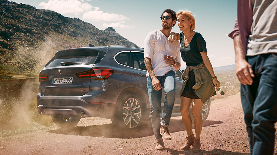 BMW X1, F48 LCI, pintura Storm Bay, perspetiva traseira a três quartos BMW X1, F48, LCI, pintura Storm Bay, perspetiva traseira a três quartos, parado numa paisagem com pessoas à frente