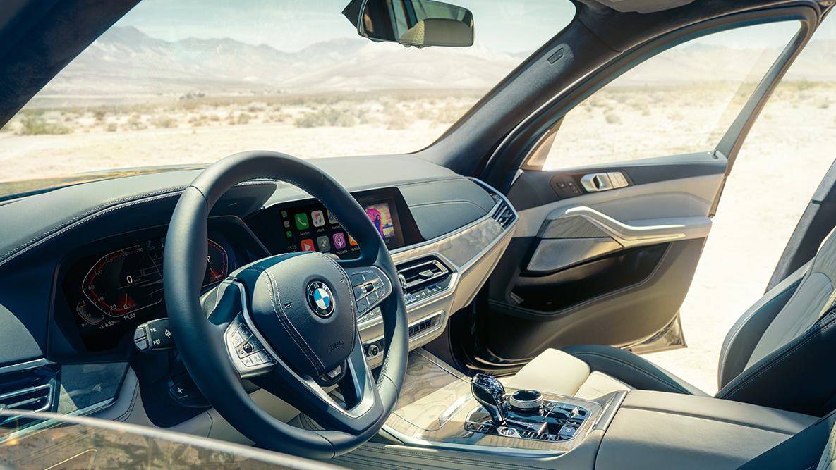 Interior do BMW X7 Interior do BMW X7: perspetiva do exterior para a primeira fila de bancos com imagem do volante, painel de instrumentos e bancos em pele
