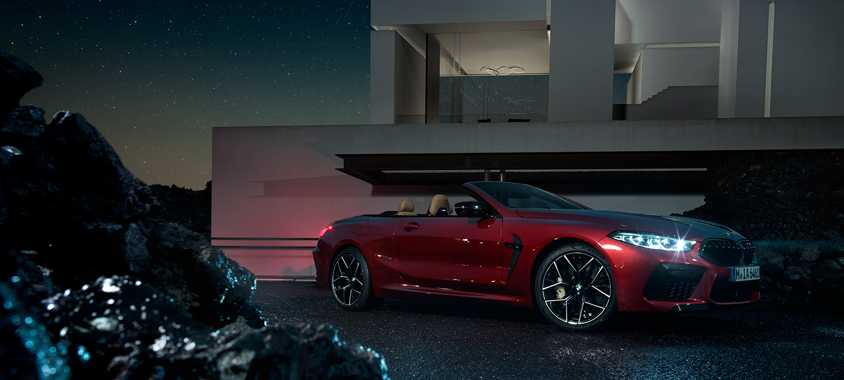 BMW M8 Competition Convertible, three-quarter side view. BMW M8 Competition Cabrio em Vermelho Motegi Metalizado, perspetiva lateral a três quartos, durante a noite em frente da vila.