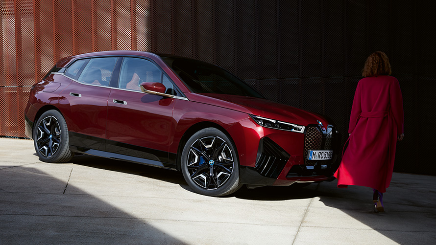 Mulher à frente do BMW iX, i20, 2021, perspetiva dianteira a três quartos Mulher com um casaco vermelho à frente do BMW iX, i20, 2021, perspetiva dianteira a três quartos