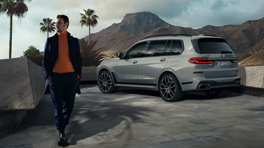 Homem à frente de um BMW X7 (G07) parado, estacionamento por controlo remoto Estacionamento por controlo remoto, homem à frente de um BMW X7 (G07) parado, perspetiva traseira a três quartos, debaixo de palmeiras com uma paisagem de montanha no fundo