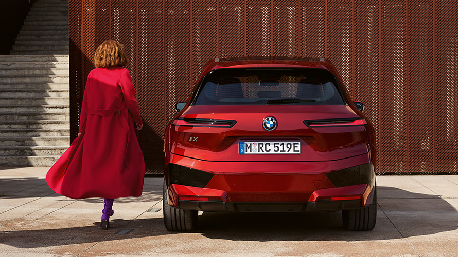 Mulher com um casaco vermelho junto ao um BMW iX (i20), fecho da porta da bagageira Fecho da porta da bagageira, mulher com um casaco vermelho junto ao um BMW iX (i20) à frente de um edifício