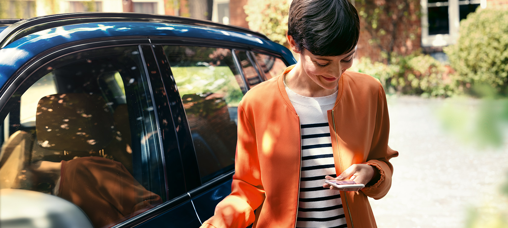 Mulher morena à frente de um BMW Mulher morena vestida com casaco cor de laranja à frente de um BMW a olhar para o smartphone.