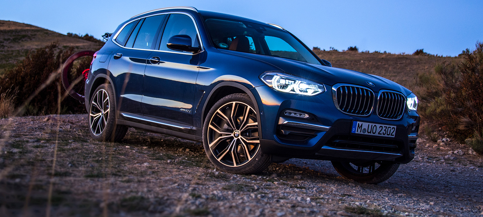 BMW X3 azul-escuro em terreno aberto BMW X3 azul-escuro estacionado com suporte para bicicletas e bicicleta de montanha em terreno aberto.