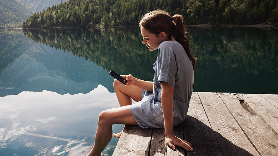Mulher sentada num pontão a olhar para o smartphone Acessórios primavera/verão BMW, mulher sentada num pontão a olhar para o smartphone