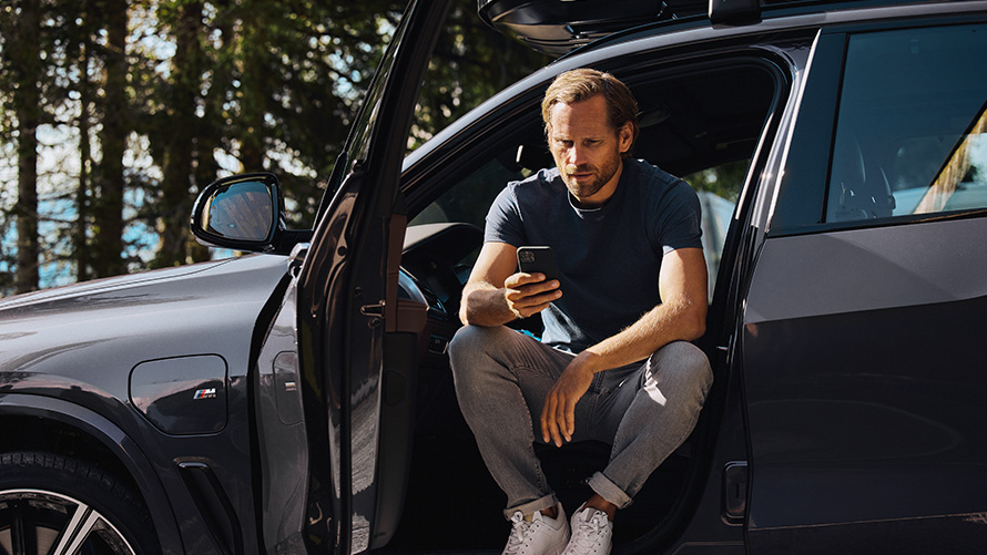 Homem sentado num BMW a olhar para o smartphone Acessórios primavera/verão BMW, homem sentado num BMW a olhar para o smartphone