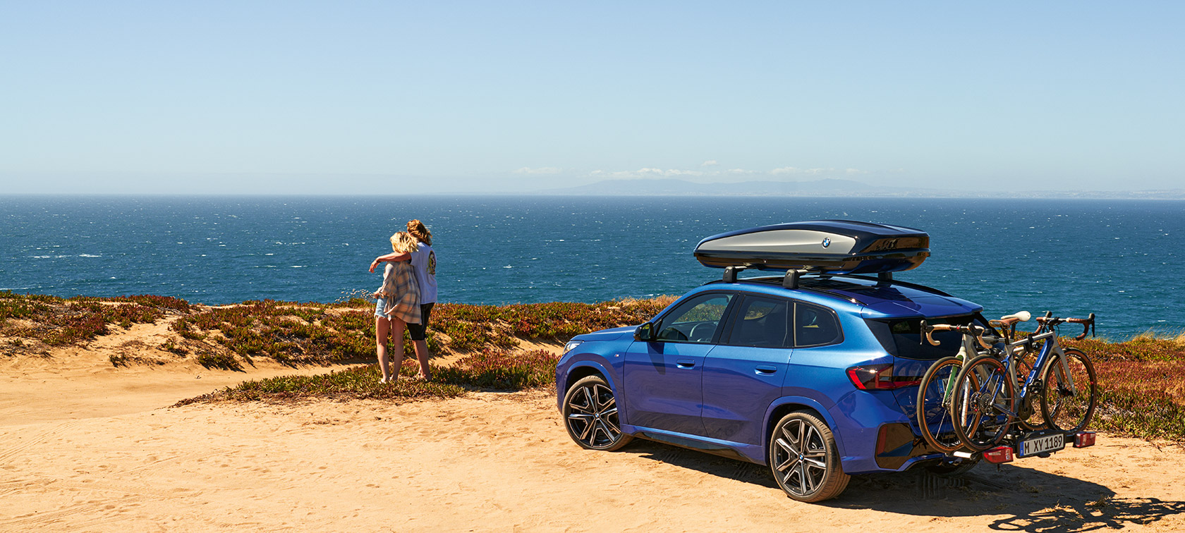 Acessórios Originais BMW para a primavera e o verão, BMW X1 U11 Acessórios Originais BMW para a primavera e o verão, BMW X1 U11, parado na areia à frente de uma costa com duas pessoas