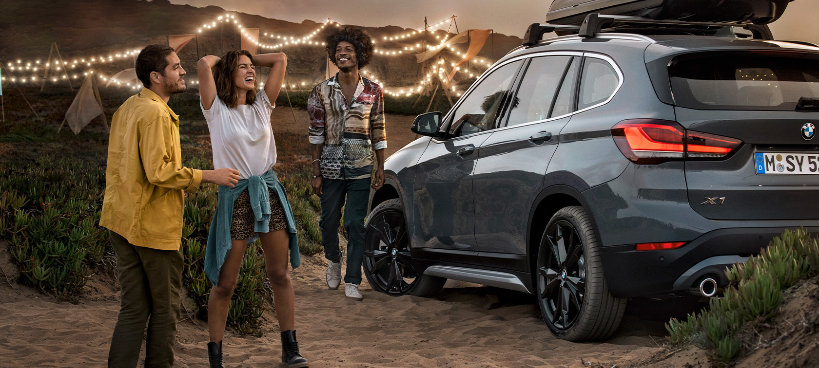 Trio animado num BMW X1 equipado com as ofertas BMW para o verão Trio feliz num BMW X1 em cinza Mineral equipado com as ofertas BMW para o verão na praia, perspetiva traseira a três quartos, equipamento BMW