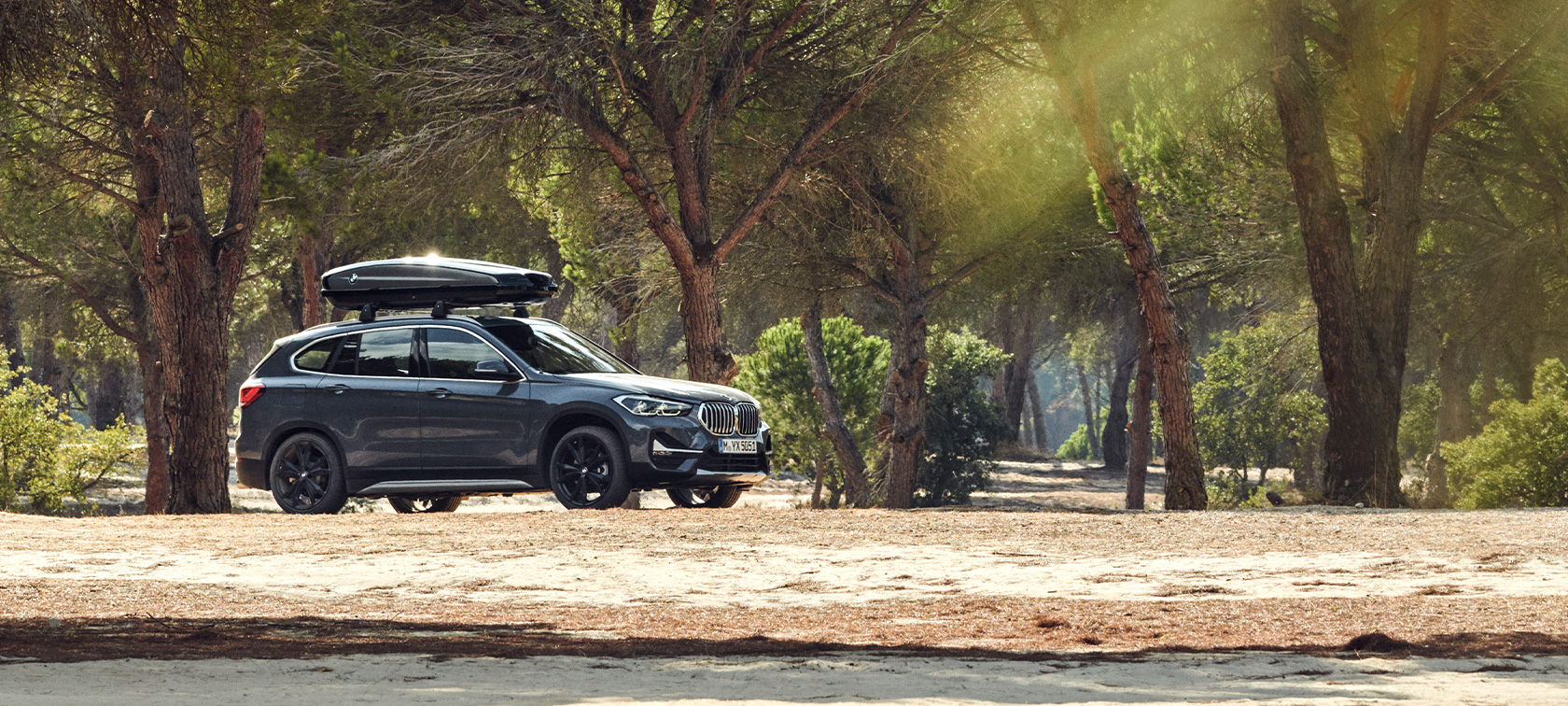 Perspetiva lateral do BMW X1 equipado com a bagageira de tejadilho BMW, ofertas BMW para o verão Perspetiva lateral do BMW X1 em cinza Mineral parado na floresta e equipado com a bagageira de tejadilho BMW, ofertas BMW para o verão