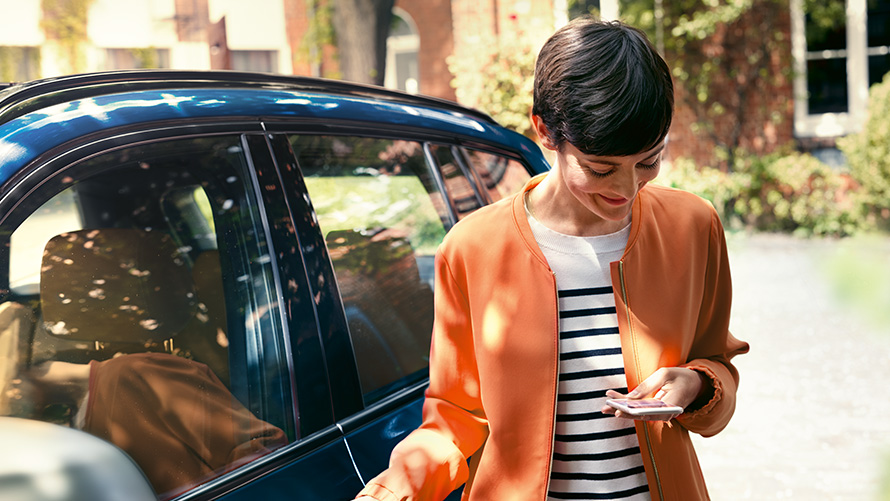 Ofertas BMW para o verão, BMW TeleServices Ofertas BMW para o verão, BMW TeleServices, uma mulher parada à frente de um BMW a utilizar o smartphone