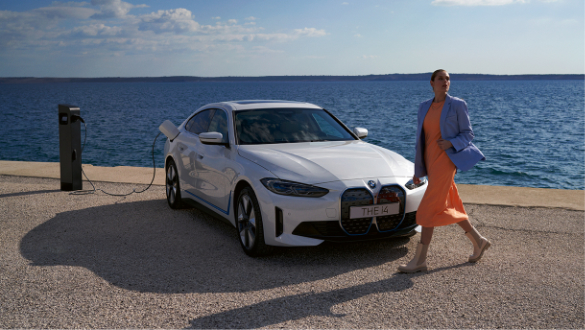 BMW i4, de cor branca, a carregar em público, enquanto mulher de vestido laranja e casaco de ganga caminha junto ao carro.