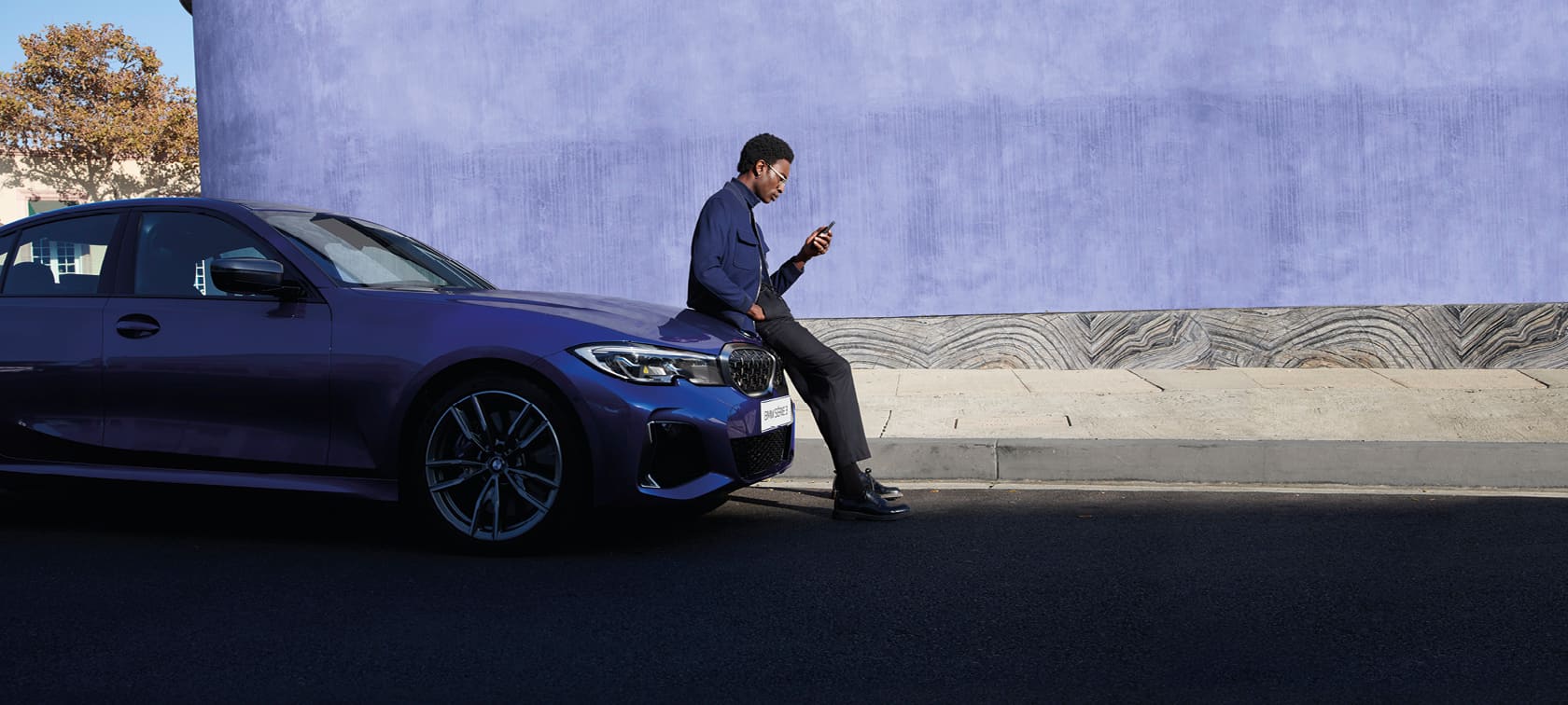Homem a consultar smartphone e sentado sobre o capô do seu BMW estacionado na rua.