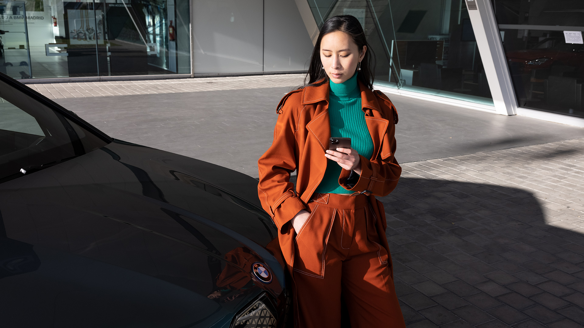 Mulher vestida de laranja ao lado de um BMW à frente de um edifício de escritórios a olhar para o seu smartphone
