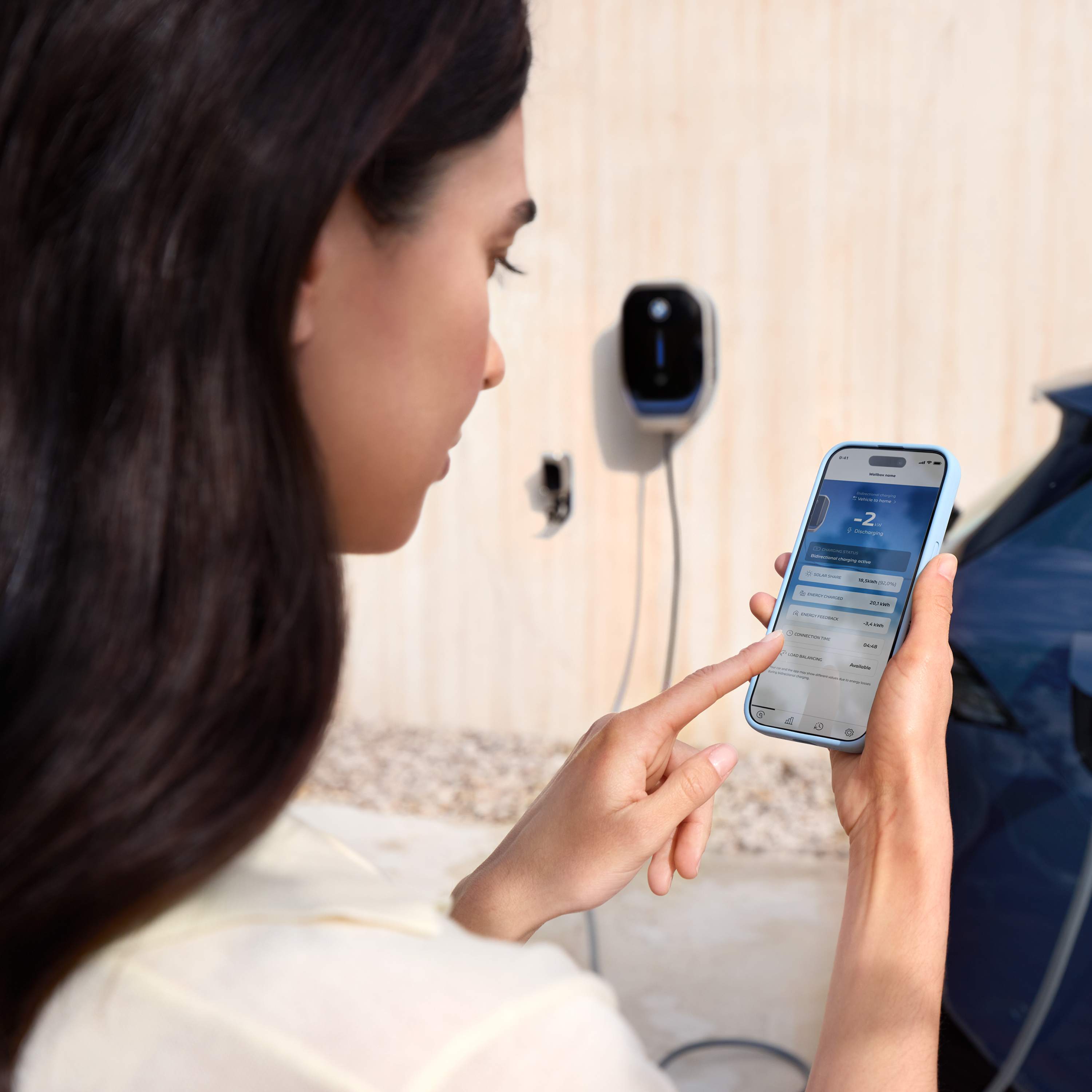 Uma mulher está a olhar para o seu smartphone, uma wallbox é visível em segundo plano